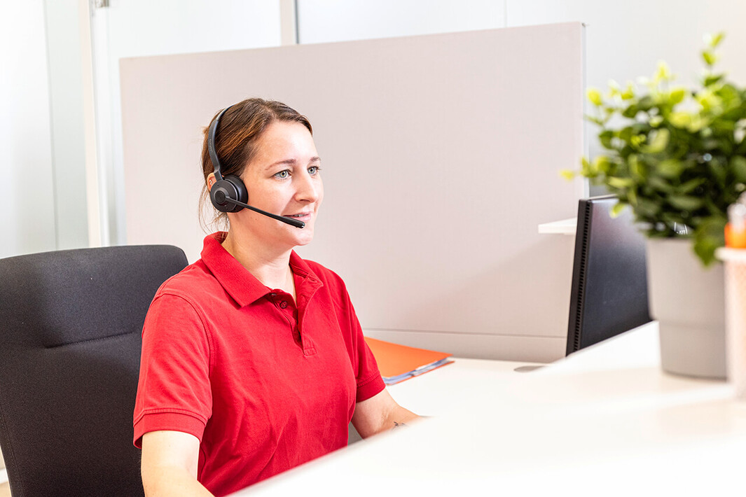 Mitarbeiterin am Empfang mit Headset und rotem T-Shirt gibt telefonische Auskunft.